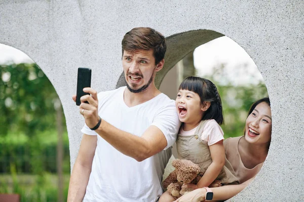 Młody Mężczyzna Robi Śmieszną Minę Podczas Fotografowania Żoną Córeczką Parku — Zdjęcie stockowe