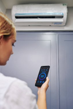 Klimada sıcaklık ayarlanırken akıllı telefonda uygulama kullanan genç bir kadın