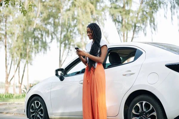 快乐的年轻黑人女人 留着长长的头发 站在她的汽车旁边 给朋友们发短信 — 图库照片