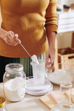 Plastik kavanoza soda tozu koyan ve sabun karışımı yaparken elektronik ölçekte ölçen bir kadın görüntüsü.