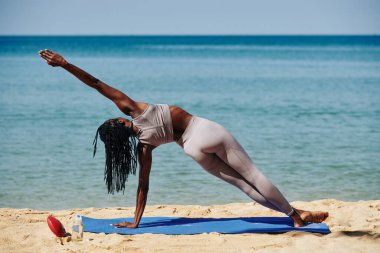 Siyah bir kadın, plajda yoga minderinin üzerinde uzun bir yan kalas yapıyor.