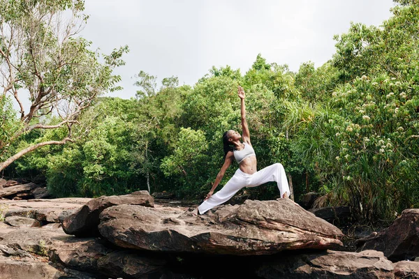 Fit Black woman practicing yoga on big rock and doing warrior pose