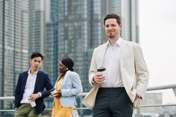 自信に満ちた若い起業家を笑顔にし 屋外に立つコーヒーを飲みながら 同僚たちは背景で話しています — ストック写真