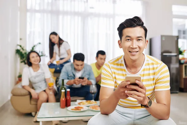 笑顔ベトナムの若い男のテキストメッセージとカメラを見るとき彼の友人食べると家のパーティーでバックグラウンドで飲む — ストック写真