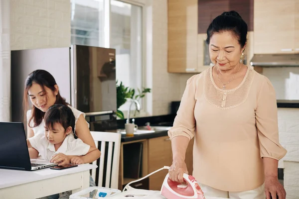 年轻的亚洲女人和女儿一起在电脑上打圈 而她的大妈则在熨烫衣服 — 图库照片