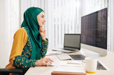 Geleneksel elbiseli gülümseyen müslüman genç kadın bilişim şirketi için yeni bir proje üzerinde çalışırken eğleniyor.
