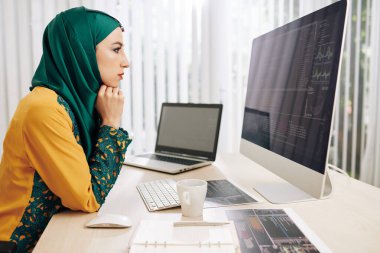 Frowning serious female muslim coder looking for mistakes in her programming code on computer screen clipart