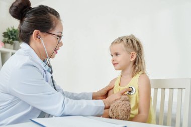 Gülümseyen olgun Vietnamlı pratisyen doktor küçük kızın kalp atışlarını dinliyor.