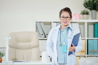 Ofisinde elinde klasörle kameraya gülümseyen olgun Asyalı doktorun portresi.