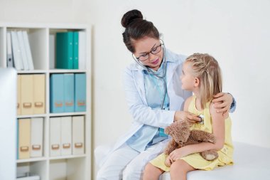Küçük kız, çocuk doktoruna bakıyor. Ona sarılıyor, kalp atışlarını ve nefesini steteskopla kontrol ediyor.