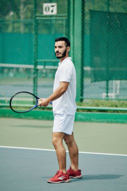 Beyaz spor elbiseli yakışıklı genç adam dışarıda tenis oynuyor.
