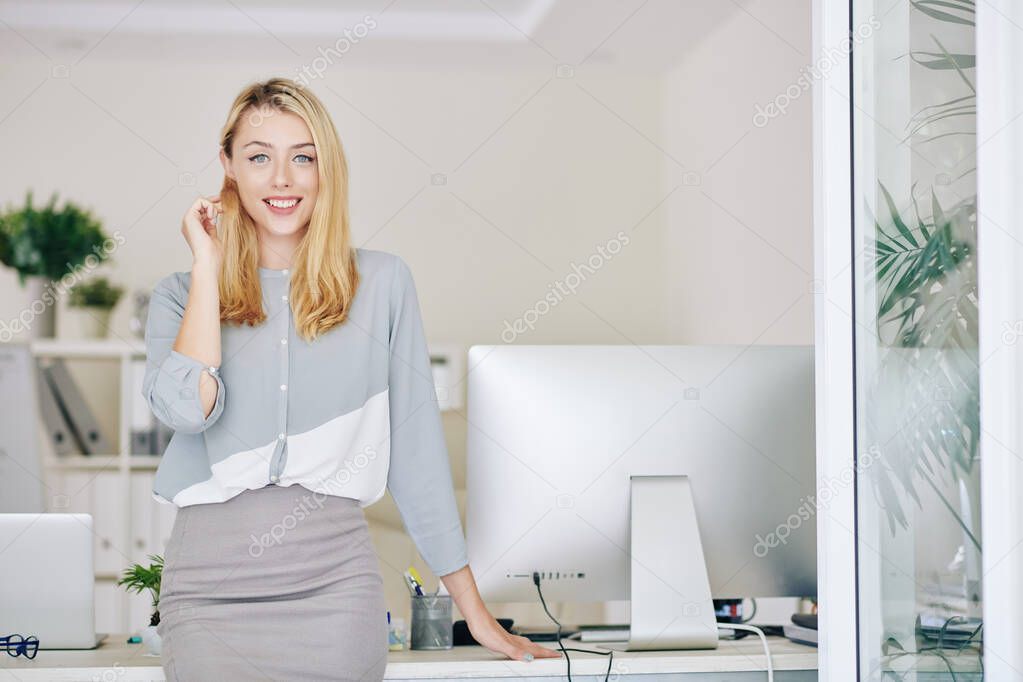 Beautiful smiling young female entrepreneur leaning on her office table and looking at camera