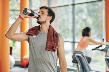 Spor salonundaki eğitimden sonra omzunda havluyla susamış bir sporcu su içiyor.