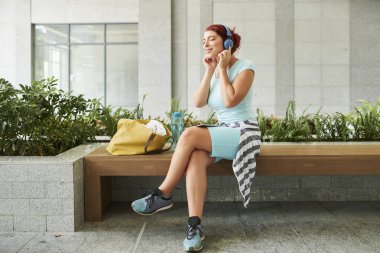 Mutlu genç kadın bankta oturuyor ve kulaklıkla müzik dinlerken zevkle gözlerini kapıyor.