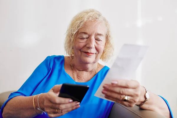 笑顔シニア女性はソファに座って モバイルアプリケーションを介して請求書を支払う — ストック写真