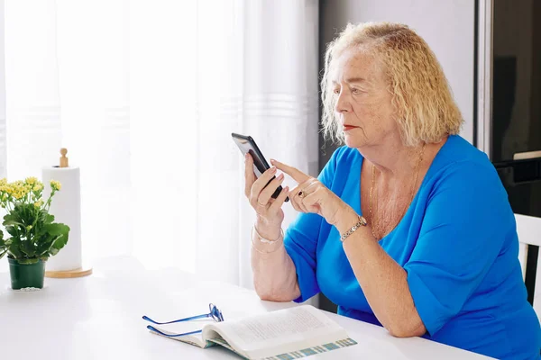 高齢女性座っているテーブルに家とチェックテキストメッセージで彼女の携帯電話 — ストック写真