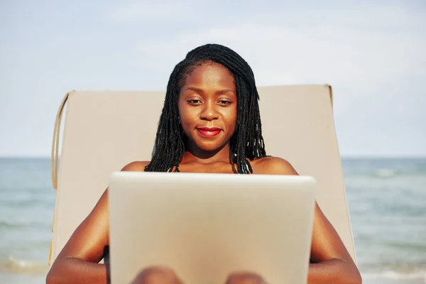 年轻漂亮的黑人妇女在躺椅上休息 在海滩上的笔记本电脑上工作的画像 — 图库照片