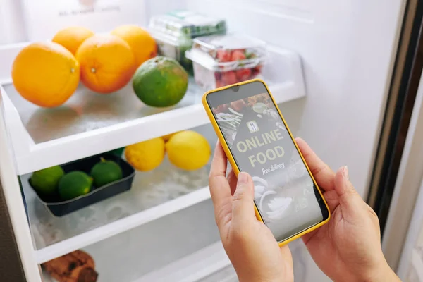 Close-up image of housewife opening refrigerator when observing what ordering products via food delivery application on smartphone