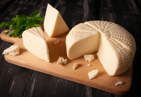 Sýr Suluguni se krájí na velké kousky. Na lehkou dřevěnou desku. Zdobené zelení. Tmavé dřevěné pozadí. — Stock fotografie