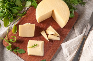 Caciotta peyniri dilimlenmiş. Dikdörtgen bir tahta üzerinde. Roka, maydanoz ve kişniş ile süslenmiş. Arka plan gri keten kumaşı. Yukarıdan görüntüle