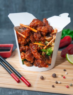 Wok için boks. Tatlı, ekşi soslu ve sebzeli domuz eti. Havuç, mantar ve brokoli. Tava kutusu kesildi. Yemek çubukları, baharatlar ve soya sosunun yanında duran bir paspasın üzerinde..