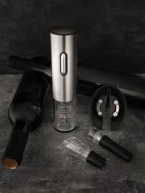 Metalik elektronik kablosuz tirbuşon. Yakınlarda iki şişe şarap, bir havalandırma ve bir vakum mantarı var. Koyu arkaplan.