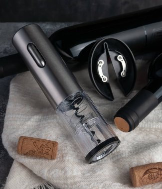 Metalik elektronik kablosuz tirbuşon. Yakınlarda iki şişe şarap ve mantar var. Ahşap ve kumaştan oluşan bir arka plan. Yakın plan..