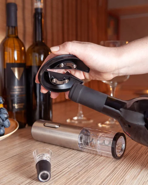 Ένα θηλυκό χέρι κρατά μια στάση για ένα τιρμπουσόν. Μεταλλικό γκρι ηλεκτρικό τιρμπουσόν και αεροβόλο βρίσκονται στο τραπέζι. Στο παρασκήνιο είναι μπουκάλια κρασί, ποτήρια και σταφύλια — Φωτογραφία Αρχείου