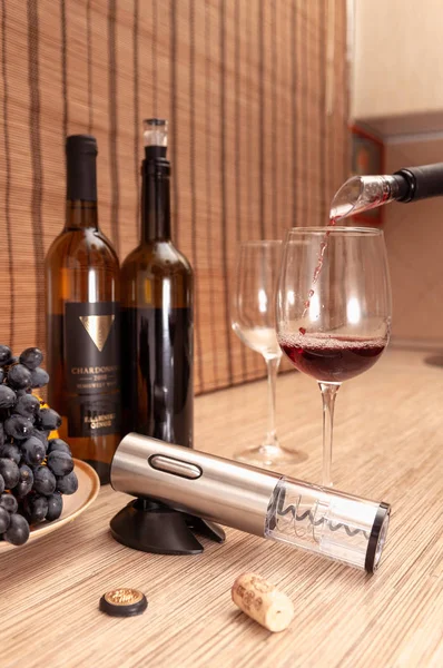 A gray metallic electric corkscrew lies on a stand. Wine is poured into the glass through the aerator. In the background are bottles of wine, glasses and grapes.