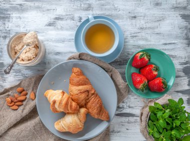 Kahvaltıda. Bir fincan çay, taze kruvasan, mis kokulu çilek ve hindistan cevizi ezmesiyle. Gri ahşap arka planda kompozisyon. Yukarıdan görüntüle.