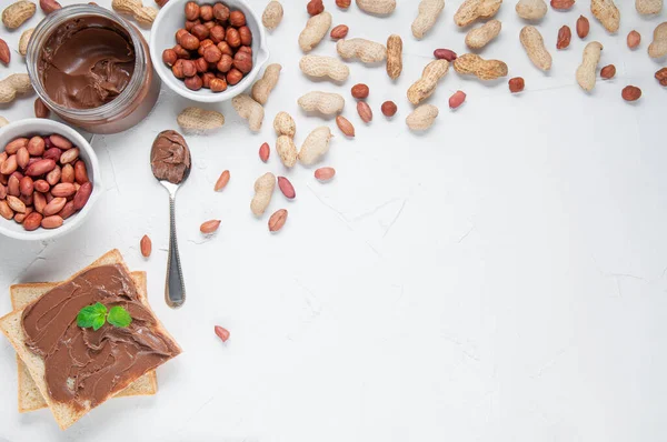 Jar of chocolate paste. Decorated with peanuts and hazelnuts. Next to a can of toast and a spoon.Flat lay composition. View from above. White background. Space for text.