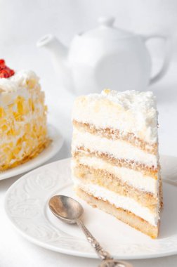 Krem şanti ve böğürtlenli sünger keki. Beyaz çikolata parçalarıyla süslenmiş. Ev yapımı. Beyaz bir tabakta. Arka planda bir kek, bir çaydanlık ve beyaz bir kupa var. Beyaz arka plan. Yakın plan..