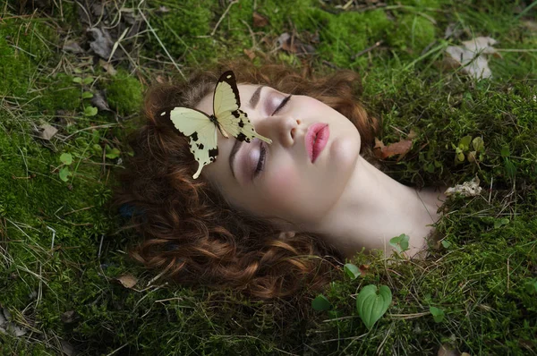 close-up portrait of cheerful young woman with closed eyes and curly hair lying in green grass with beautiful butterfly on face