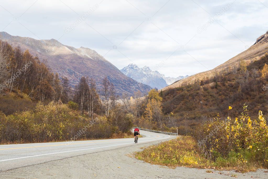 A cyclist biking down a road in the Alaskan mountains in autumn. Clouds overhead, double yellow line in center.