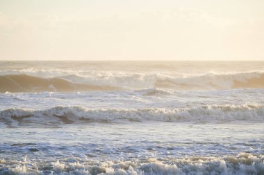  Okyanustaki su dalgaları sahile doğru yuvarlanıyor. Sarı bir pus arka planda bulutlar ile havai.