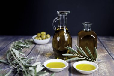 İki yağ şişesi, bir avuç yeşil zeytin kabı, zeytin dalı ve ahşap masa ve siyah arka planda iki yağ kasesi.