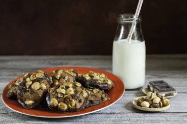 Kırmızı tabakta fındık ve çikolata parçalı kurabiyeler, fındık, çikolata parçaları ve ahşap bir masada pipetle birlikte bir şişe süt ve fotokopi alanı olan koyu kahverengi arka plan.