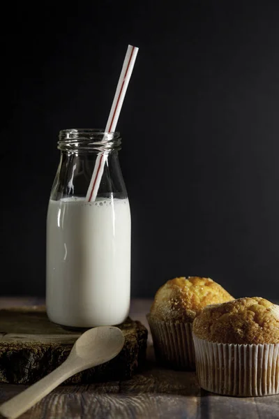 Fresh milk in a glass bottle, muffins and wooden spoon, on a wooden table and black backgroundl. The concept of healthy organic, farm products. Rustic style of still life. Copy space for text.