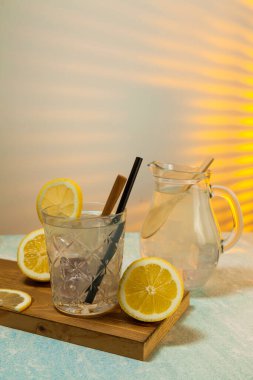 Kesme tahtasına buz, kaşık ve saman konmuş limonata bardağı, limonata sürahisi ve arka pencereden içeri güneş ışığı girerken arka planda limonlar. Boşluğu kopyala