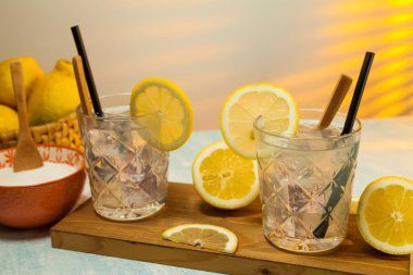 Kesme tahtasının üzerinde buz, kaşık ve saman bulunan iki bardak limonata, limon dilimleri, limonlu sepet ve arka planda şeker olan arka pencereden giren güneş ışığıyla dolu bir kase. Boşluğu kopyala