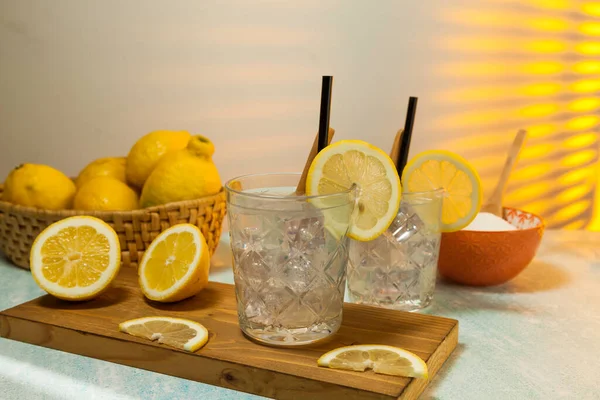 two glasses of lemonade with ice, spoon and straw on cutting board, cut lemons, basket with lemons and bowl with sugar on background with sunlight entering through rear window. Copy space