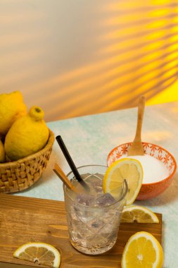 limonata bardağı, buz, kaşık ve kamış kesme tahtasına, limon dilimlemeye, limonlu sepete ve arka planda şekerli kaseye güneş ışığı arka pencereden giriyor. Boşluğu kopyala