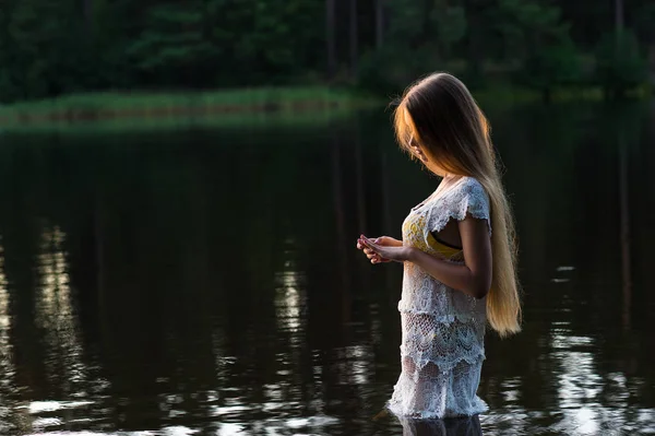 Encantadora joven en vestido blanco de pie en el agua en la puesta del sol —  Fotos de Stock