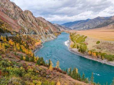 River running through the mountains. Dark turquoise flow of Katun River and surrounding mountains along the Chuysky Trakt road in the Altai Mountains region of Siberia. Yellow larches and other autumn trees in front on a cloudy day. clipart