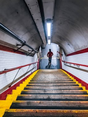 Londra Metrosu, merdivenlerden aşağı yürüme. Tüp Altgeçit merdivenlerden acele kırmızı ceketli genç adam. Beyaz seramik duvar karoları ve tebeşir çiftlik metro istasyonunda kırmızı Küpeşte ile metal merdiven.