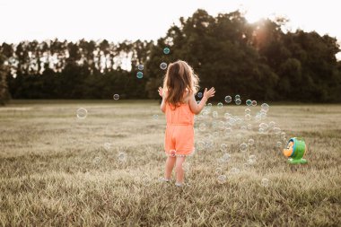 Gün batımında tarlada sabun köpüğü yakalayan turuncu tulumlu küçük kız..