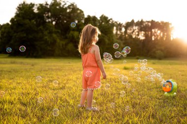 Gün batımında tarlada sabun köpüğü yakalayan turuncu tulumlu küçük kız..