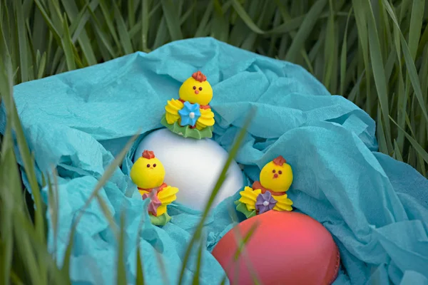 Three toy chickens and three easter eggs on the grass in a blue basket