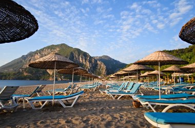 Plaj şemsiyeleri ve güneş şezlongları sabahın köründe Cirali sahilinde, insan olmadan. Arka planda dağlar ve mavi gökyüzü. Güneşli bir gün. Antalya, Türkiye.