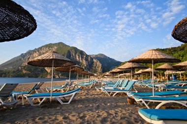 Plaj şemsiyeleri ve güneş şezlongları sabahın köründe Cirali sahilinde, insan olmadan. Arka planda dağlar ve mavi gökyüzü. Güneşli bir gün. Antalya, Türkiye.